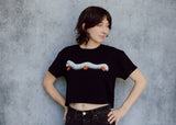 A model wearing a black cropped t-shirt featuring a millipede in a ghost costume carrying jack o'lantern candy pails. 