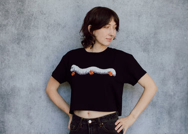 A model wearing a black cropped t-shirt featuring a millipede in a ghost costume carrying jack o'lantern candy pails. 