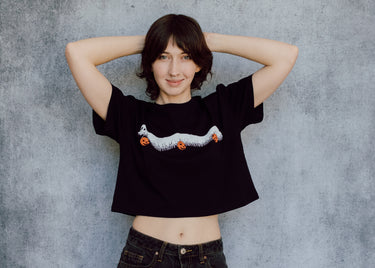 A model wearing a black cropped t-shirt featuring a millipede in a ghost costume carrying jack o'lantern candy pails. 