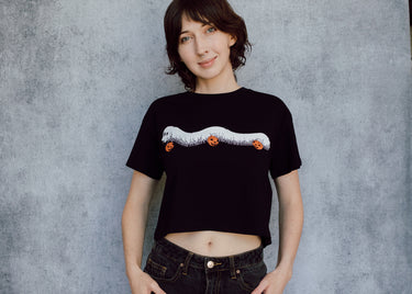 A model wearing a black cropped t-shirt featuring a millipede in a ghost costume carrying jack o'lantern candy pails. 
