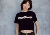 A model wearing a black cropped t-shirt featuring a millipede in a ghost costume carrying jack o'lantern candy pails. 