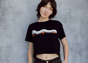 A model wearing a black cropped t-shirt featuring a millipede in a ghost costume carrying jack o'lantern candy pails. 