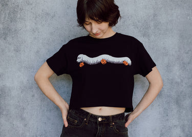 A model wearing a black cropped t-shirt featuring a millipede in a ghost costume carrying jack o'lantern candy pails. 