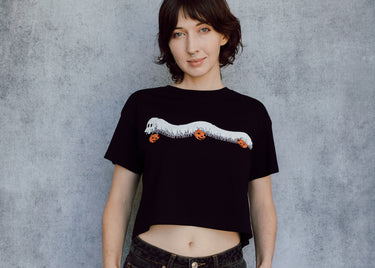 A model wearing a black cropped t-shirt featuring a millipede in a ghost costume carrying jack o'lantern candy pails. 