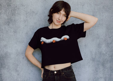 A model wearing a black cropped t-shirt featuring a millipede in a ghost costume carrying jack o'lantern candy pails. 