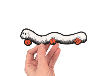A hand holding a die cut sticker of a millipede in a ghost costumer carrying three jack 'o lantern candy pails. 
