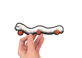 A hand holding a die cut sticker of a millipede in a ghost costumer carrying three jack 'o lantern candy pails. 