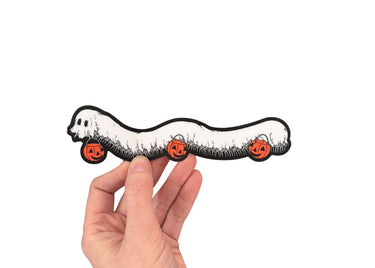 A hand holding a die cut sticker of a millipede in a ghost costumer carrying three jack 'o lantern candy pails. 