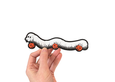 A hand holding a die cut sticker of a millipede in a ghost costumer carrying three jack 'o lantern candy pails. 