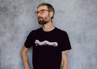 A model wearing a black t-shirt with a hand drawn illustration of a millipede in a ghost costume carrying jack'o lantern candy pails. 