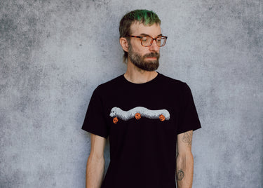 A model wearing a black t-shirt with a hand drawn illustration of a millipede in a ghost costume carrying jack'o lantern candy pails. 