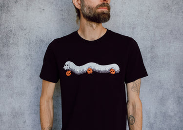 A model wearing a black t-shirt with a hand drawn illustration of a millipede in a ghost costume carrying jack'o lantern candy pails. 
