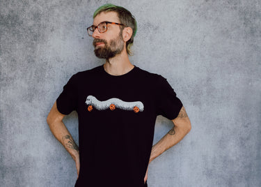 A model wearing a black t-shirt with a hand drawn illustration of a millipede in a ghost costume carrying jack'o lantern candy pails. 
