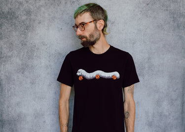 A model wearing a black t-shirt with a hand drawn illustration of a millipede in a ghost costume carrying jack'o lantern candy pails. 