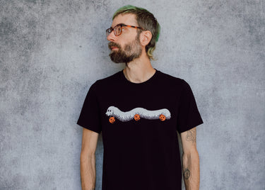 A model wearing a black t-shirt with a hand drawn illustration of a millipede in a ghost costume carrying jack'o lantern candy pails. 