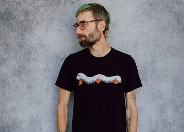 A model wearing a black t-shirt with a hand drawn illustration of a millipede in a ghost costume carrying jack'o lantern candy pails. 