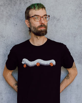 A model wearing a black t-shirt with a hand drawn illustration of a millipede in a ghost costume carrying jack'o lantern candy pails. 