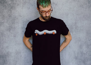 A model wearing a black t-shirt with a hand drawn illustration of a millipede in a ghost costume carrying jack'o lantern candy pails. 