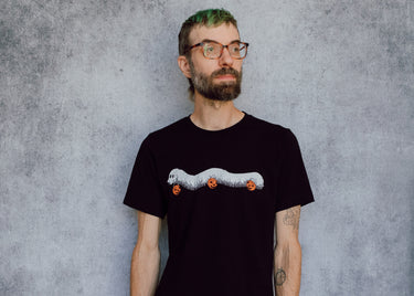 A model wearing a black t-shirt with a hand drawn illustration of a millipede in a ghost costume carrying jack'o lantern candy pails. 