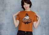 A model wearing an orange cropped t-shirt featuring a toad with a witch hat in a pumpkin patch printed in cream and black ink. 