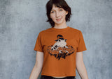 A model wearing an orange cropped t-shirt featuring a toad with a witch hat in a pumpkin patch printed in cream and black ink. 