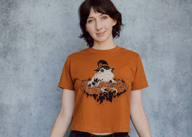 A model wearing an orange cropped t-shirt featuring a toad with a witch hat in a pumpkin patch printed in cream and black ink. 