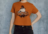 A model wearing an orange cropped t-shirt featuring a toad with a witch hat in a pumpkin patch printed in cream and black ink. 