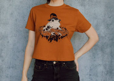 A model wearing an orange cropped t-shirt featuring a toad with a witch hat in a pumpkin patch printed in cream and black ink. 