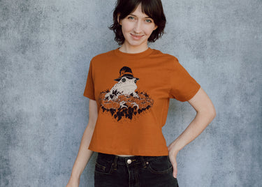 A model wearing an orange cropped t-shirt featuring a toad with a witch hat in a pumpkin patch printed in cream and black ink. 