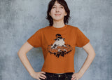 A model wearing an orange cropped t-shirt featuring a toad with a witch hat in a pumpkin patch printed in cream and black ink. 