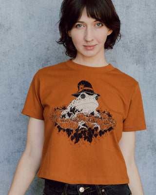 A model wearing an orange cropped t-shirt featuring a toad with a witch hat in a pumpkin patch printed in cream and black ink. 