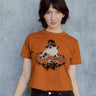 A model wearing an orange cropped t-shirt featuring a toad with a witch hat in a pumpkin patch printed in cream and black ink. 