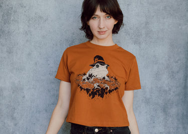 A model wearing an orange cropped t-shirt featuring a toad with a witch hat in a pumpkin patch printed in cream and black ink. 