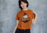 A model wearing an orange cropped t-shirt featuring a toad with a witch hat in a pumpkin patch printed in cream and black ink. 