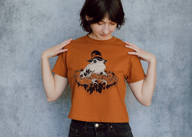 A model wearing an orange cropped t-shirt featuring a toad with a witch hat in a pumpkin patch printed in cream and black ink. 