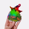 A hand holding a holographic sticker featuring a toad in a witch hat with pumpkins.
