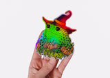 A hand holding a holographic sticker featuring a toad in a witch hat with pumpkins.
