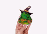 A hand holding a holographic sticker featuring a toad in a witch hat with pumpkins.
