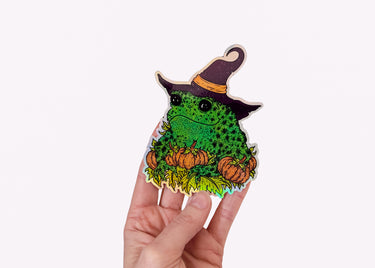 A hand holding a holographic sticker featuring a toad in a witch hat with pumpkins.