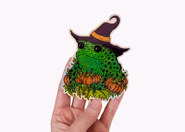 A hand holding a holographic sticker featuring a toad in a witch hat with pumpkins.