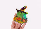 A hand holding a holographic sticker featuring a toad in a witch hat with pumpkins.
