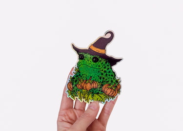 A hand holding a holographic sticker featuring a toad in a witch hat with pumpkins.