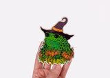 A hand holding a holographic sticker featuring a toad in a witch hat with pumpkins.