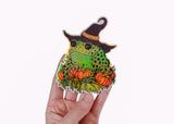 A hand holding a holographic sticker featuring a toad in a witch hat with pumpkins.