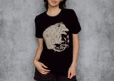 A woman with black hair is wearing a black t-shirt with a detailed cream illustration of a possum on the front, standing against a textured gray wall.