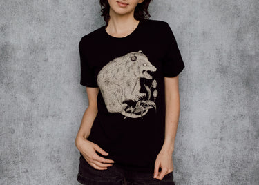 A woman with black hair is wearing a black t-shirt with a detailed cream illustration of a possum on the front, standing against a textured gray wall.