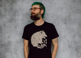A man with a beard, glasses, and bright green hair is wearing a black t-shirt with a detailed cream illustration of a possum on the front, standing against a textured gray wall.