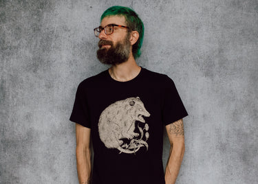 A man with a beard, glasses, and bright green hair is wearing a black t-shirt with a detailed cream illustration of a possum on the front, standing against a textured gray wall.